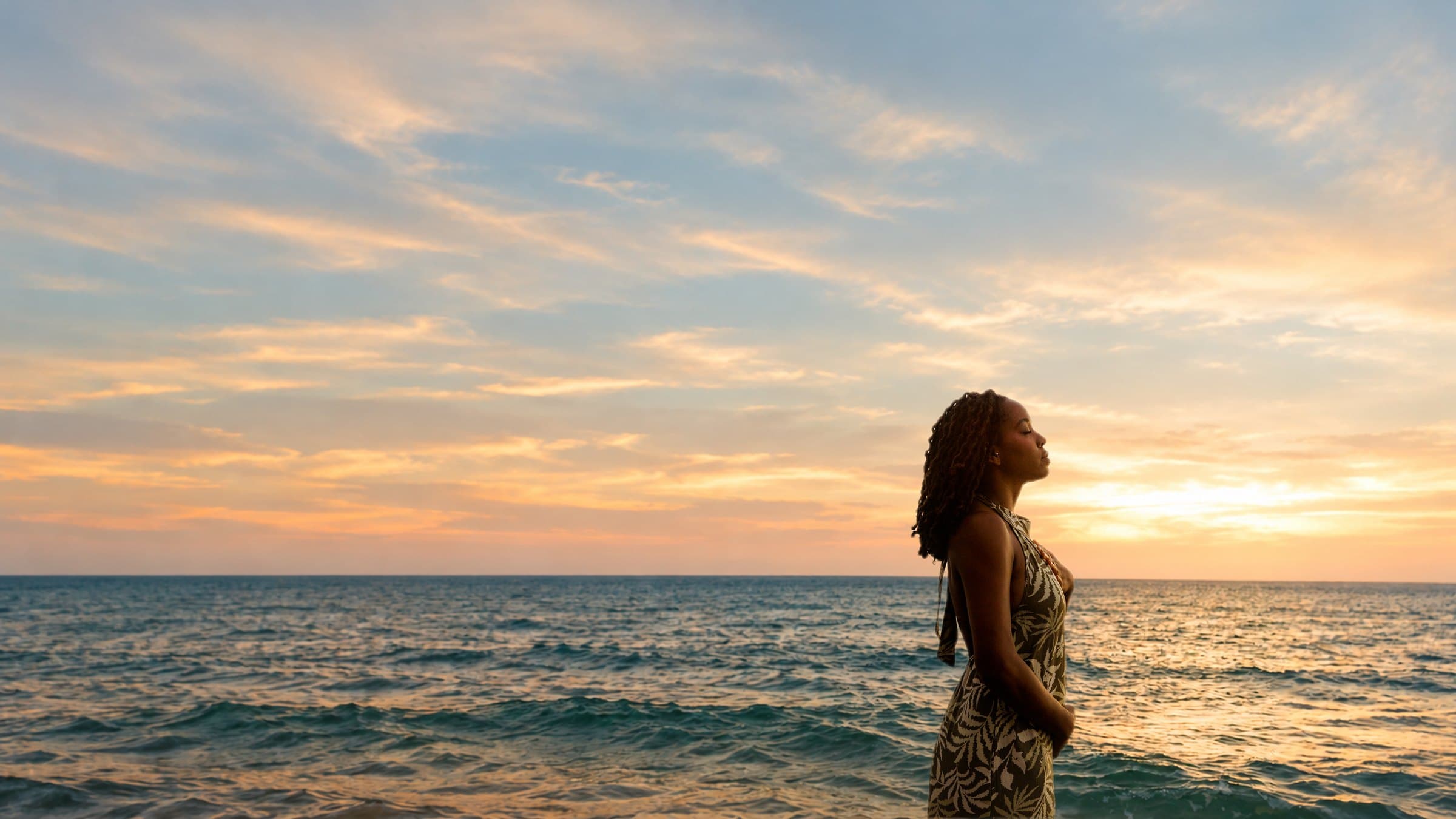 Dr. Cortney Beasley standing by the ocean at sunset, eyes closed, hand on her heart, with open sea and sky around her